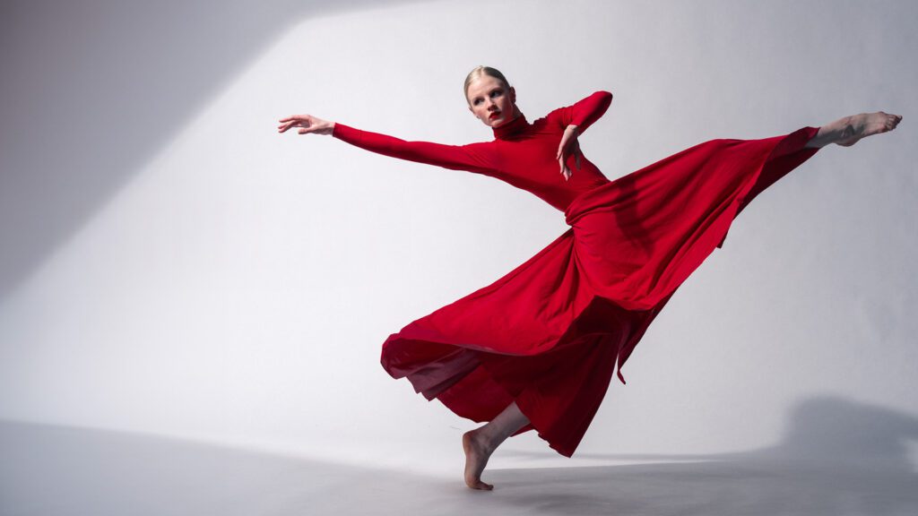 A dancer in a red dress performs a leap against a light backdrop.